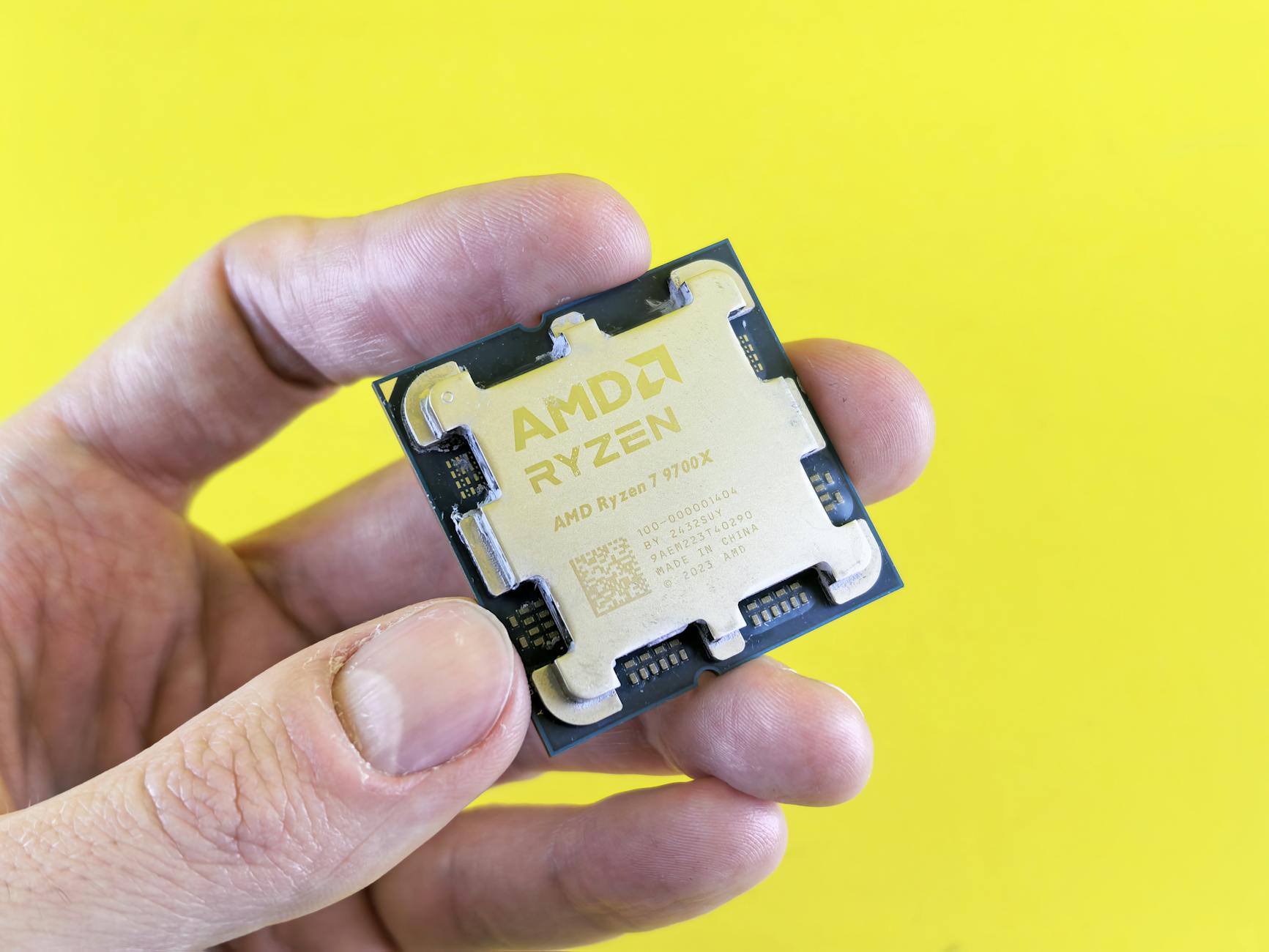 AMD Ryzen CPU held between fingers against a yellow background, showing the chip die and labeling